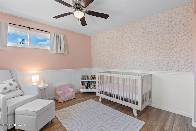 208 Pine Cone Trl, Ormond Beach, FL 32174 - bedroom with a wainscoted wall, a nursery area, wallpapered walls, wood finished floors, and a ceiling fan