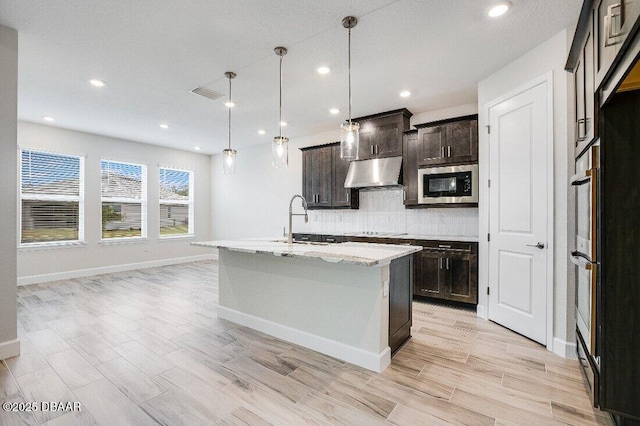 kitchen featuring pendant lighting, decorative backsplash, dark brown cabinetry, stainless steel appliances, and a center island with sink