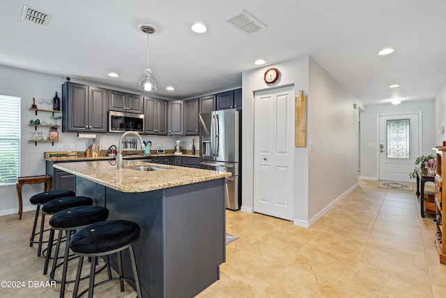 kitchen featuring stainless steel appliances, a center island with sink, sink, light stone counters, and hanging light fixtures