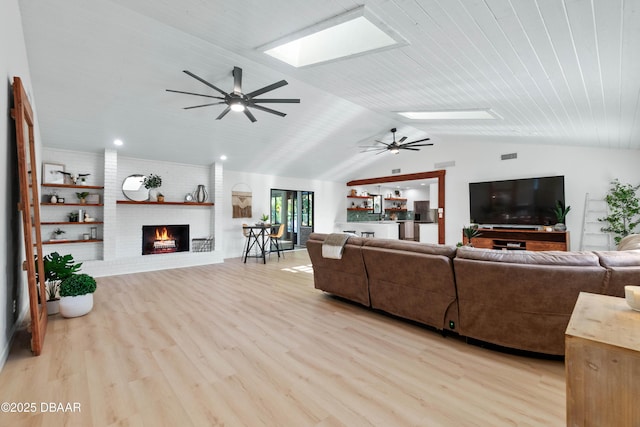 living room featuring ceiling fan, vaulted ceiling with skylight, light hardwood / wood-style floors, brick wall, and a brick fireplace