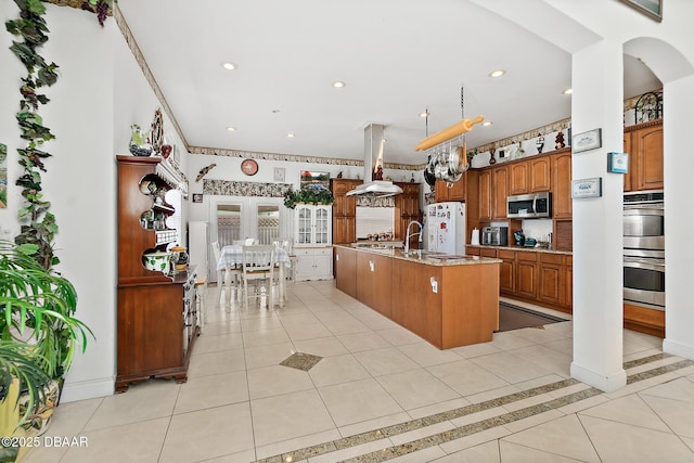 kitchen with island range hood, a kitchen island with sink, light tile patterned floors, stainless steel appliances, and light stone countertops