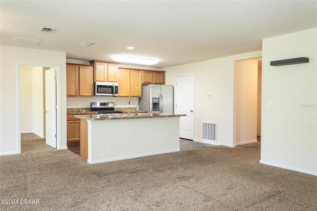 kitchen featuring dark colored carpet, a textured ceiling, a kitchen island with sink, appliances with stainless steel finishes, and dark stone countertops