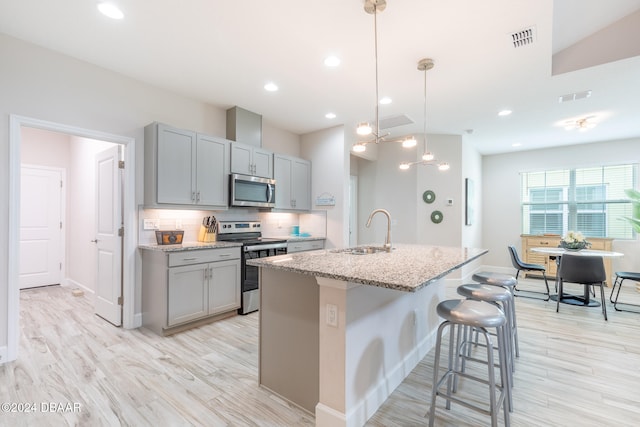 kitchen featuring stainless steel appliances, a center island with sink, sink, light stone counters, and gray cabinetry