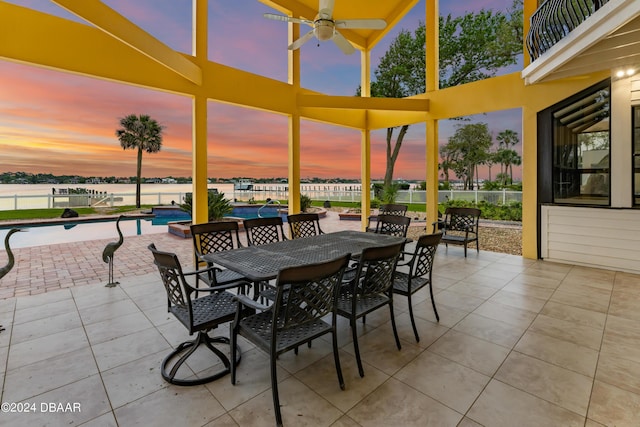 patio terrace at dusk with a fenced in pool, ceiling fan, a balcony, and a water view