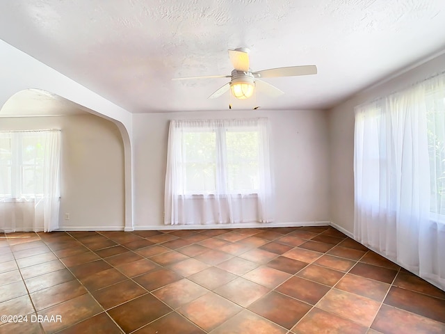 empty room featuring arched walkways, a textured ceiling, dark tile patterned floors, a ceiling fan, and baseboards