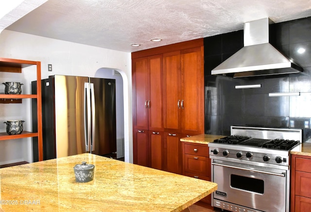 kitchen with tasteful backsplash, brown cabinetry, wall chimney exhaust hood, light stone countertops, and stainless steel appliances