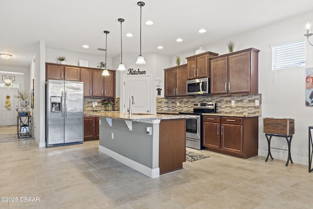 kitchen with a breakfast bar, hanging light fixtures, stainless steel appliances, light stone countertops, and an island with sink