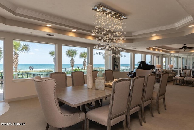 carpeted dining space with a raised ceiling, ceiling fan, ornamental molding, and a water view