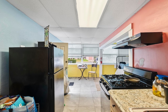 kitchen with black fridge, ventilation hood, light tile patterned flooring, a paneled ceiling, and stainless steel range with gas cooktop