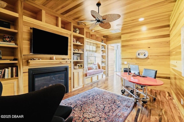 office featuring wooden walls, a ceiling fan, wood finished floors, wood ceiling, and a glass covered fireplace