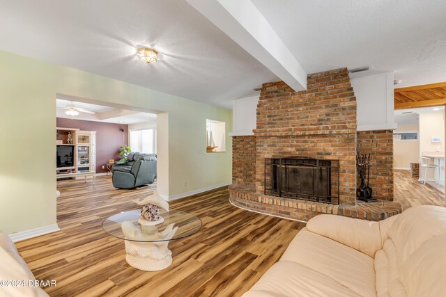 living room with ceiling fan, a brick fireplace, beamed ceiling, a textured ceiling, and light wood-type flooring