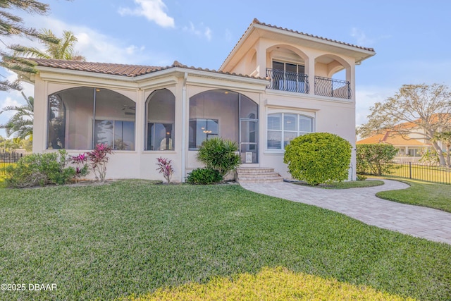 mediterranean / spanish home featuring a balcony, fence, a sunroom, stucco siding, and a front yard