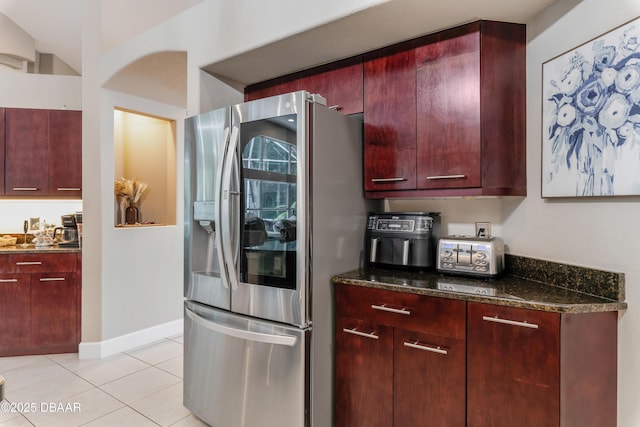 kitchen featuring stainless steel refrigerator with ice dispenser, reddish brown cabinets, light tile patterned floors, dark stone countertops, and baseboards