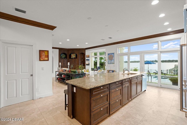 kitchen with light stone counters, crown molding, sink, an island with sink, and a water view