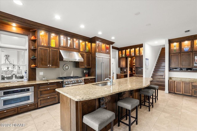 kitchen with range hood, light stone countertops, sink, high end appliances, and a kitchen island with sink