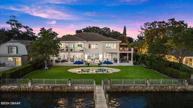 back house at dusk with a water view, a patio, a balcony, and a yard