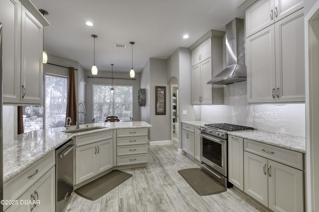 kitchen featuring kitchen peninsula, stainless steel appliances, sink, wall chimney range hood, and hanging light fixtures