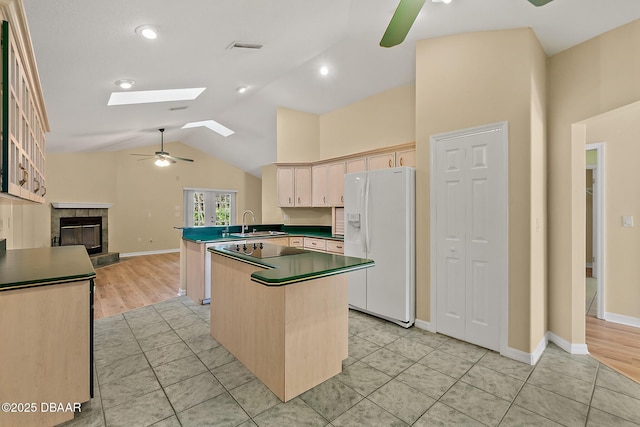 kitchen featuring ceiling fan, white refrigerator with ice dispenser, lofted ceiling with skylight, a tiled fireplace, and dark countertops