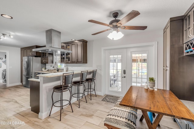 kitchen featuring a breakfast bar, island range hood, dark brown cabinetry, kitchen peninsula, and stainless steel refrigerator with ice dispenser