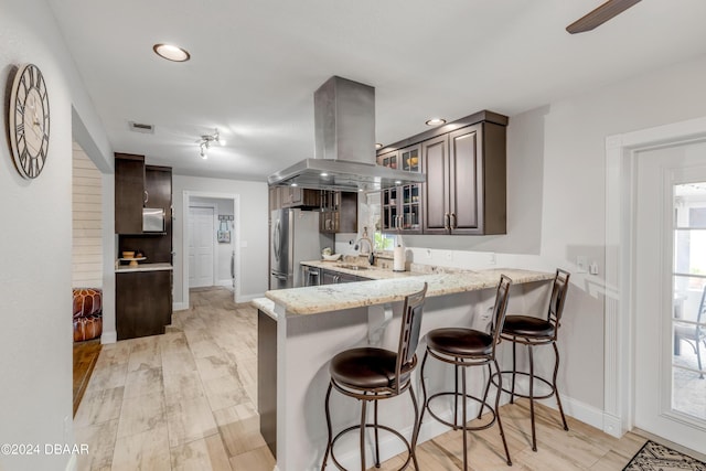 kitchen with sink, stainless steel refrigerator, dark brown cabinets, island exhaust hood, and a kitchen bar