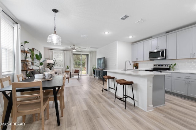 kitchen featuring gray cabinetry, backsplash, an island with sink, decorative light fixtures, and stainless steel appliances