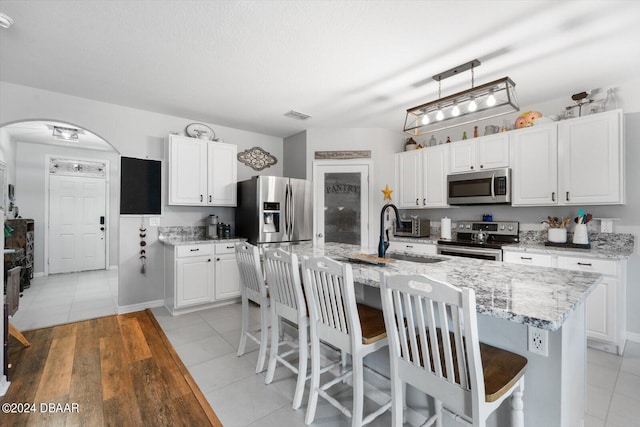 kitchen with stainless steel appliances, white cabinetry, light stone countertops, an island with sink, and light hardwood / wood-style floors