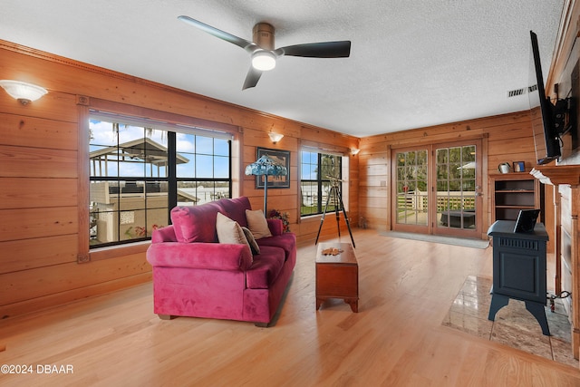 living room with a wood stove, ceiling fan, light hardwood / wood-style floors, a textured ceiling, and wooden walls