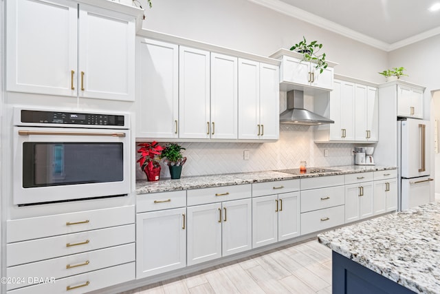 kitchen featuring white appliances, wall chimney exhaust hood, decorative backsplash, ornamental molding, and white cabinetry