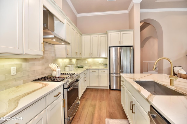 kitchen featuring crown molding, wall chimney range hood, stainless steel appliances, white cabinetry, and a sink