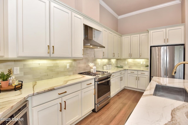 kitchen with ornamental molding, a sink, appliances with stainless steel finishes, white cabinetry, and wall chimney exhaust hood