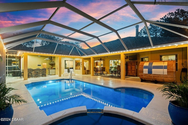 pool at dusk with an outdoor bar, a patio area, ceiling fan, and glass enclosure