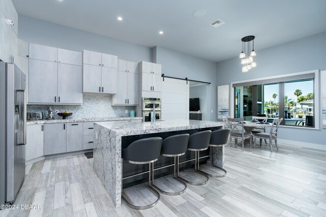 kitchen with appliances with stainless steel finishes, light stone countertops, an island with sink, a barn door, and pendant lighting