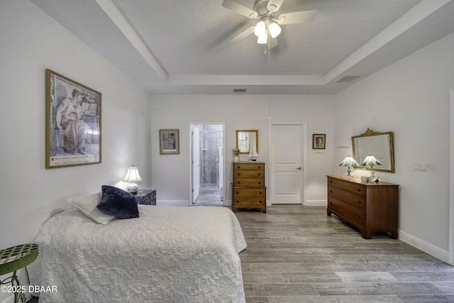 bedroom with visible vents, wood finished floors, baseboards, ensuite bathroom, and a tray ceiling