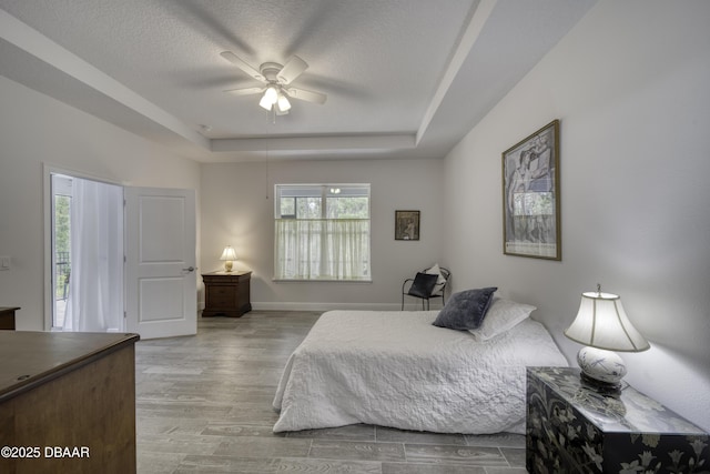 bedroom with ceiling fan, a textured ceiling, wood finished floors, baseboards, and a tray ceiling
