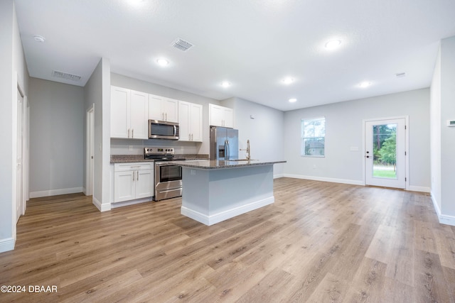 kitchen featuring stainless steel appliances, white cabinetry, a center island with sink, dark stone counters, and light wood-type flooring