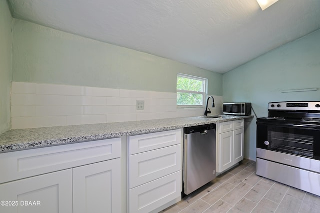 kitchen with vaulted ceiling, appliances with stainless steel finishes, white cabinetry, sink, and light stone countertops