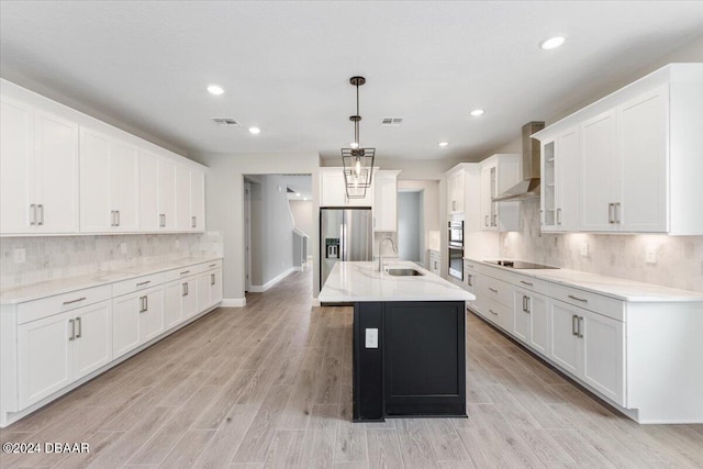 kitchen with wall chimney range hood, sink, pendant lighting, white cabinetry, and an island with sink