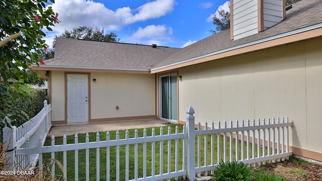 view of exterior entry with a patio, a lawn, fence, and roof with shingles