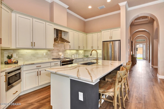 kitchen with a center island with sink, wall chimney exhaust hood, light wood-type flooring, light stone counters, and stainless steel appliances