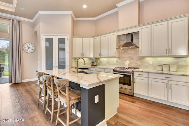 kitchen with a kitchen island with sink, wall chimney range hood, stainless steel gas stove, light wood-type flooring, and light stone counters