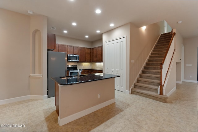 kitchen with stainless steel appliances, a kitchen island with sink, light tile patterned flooring, backsplash, and dark stone countertops