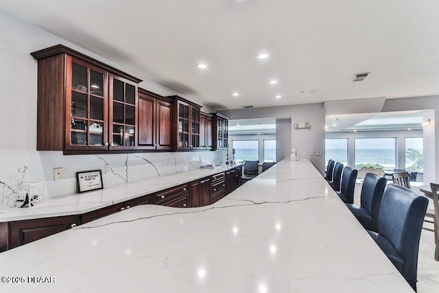 kitchen featuring decorative backsplash, glass insert cabinets, light stone counters, a kitchen bar, and recessed lighting