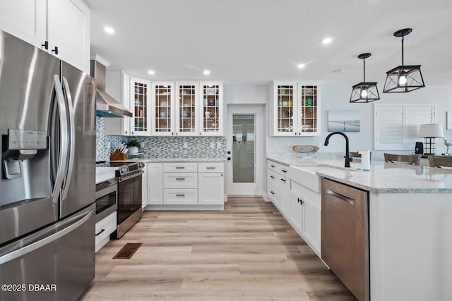 kitchen featuring light stone countertops, appliances with stainless steel finishes, wall chimney range hood, decorative light fixtures, and white cabinets