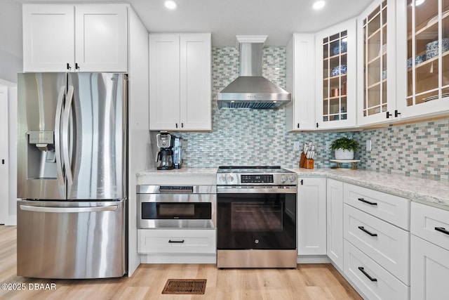 kitchen featuring appliances with stainless steel finishes, white cabinetry, wall chimney range hood, backsplash, and light stone counters