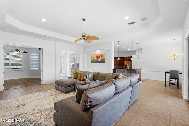 living room with a wealth of natural light, ceiling fan with notable chandelier, and a tray ceiling