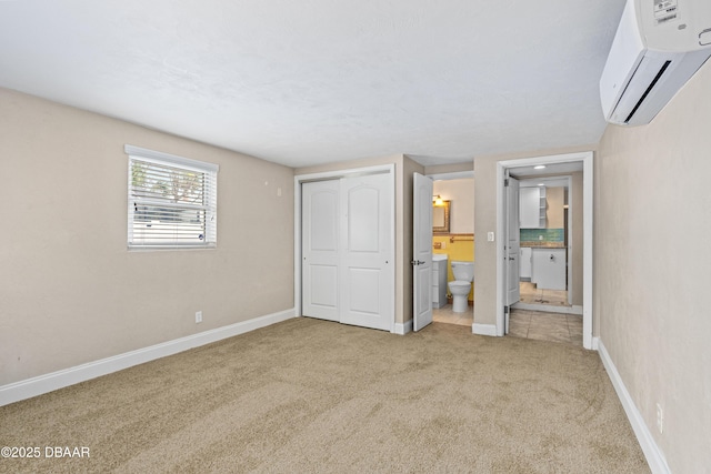 unfurnished bedroom with light colored carpet, baseboards, a closet, a wall mounted air conditioner, and ensuite bath