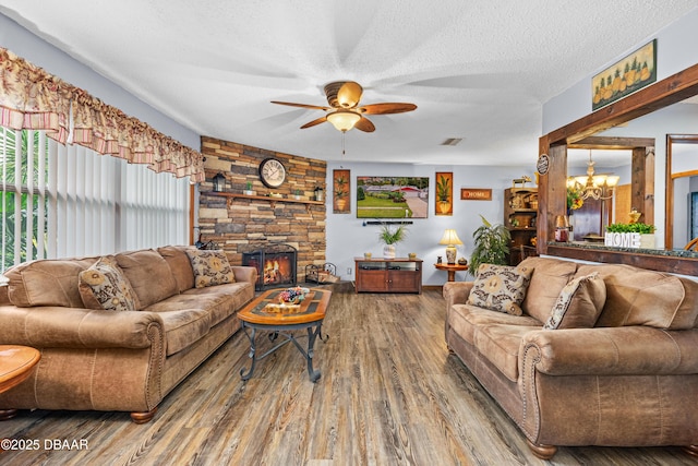 living room with a textured ceiling, ceiling fan with notable chandelier, wood-type flooring, and a stone fireplace