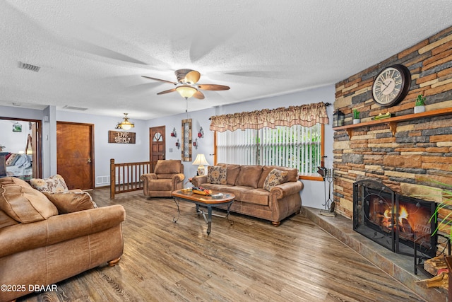 living room with ceiling fan, a textured ceiling, wood-type flooring, and a stone fireplace