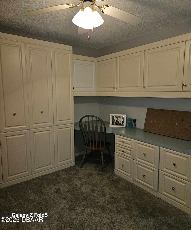 unfurnished office featuring ceiling fan, built in desk, a textured ceiling, and dark colored carpet