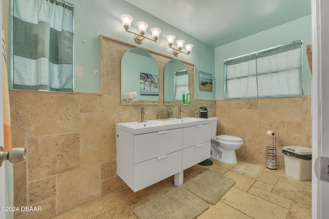 full bath with toilet, a sink, tile walls, double vanity, and stone tile flooring